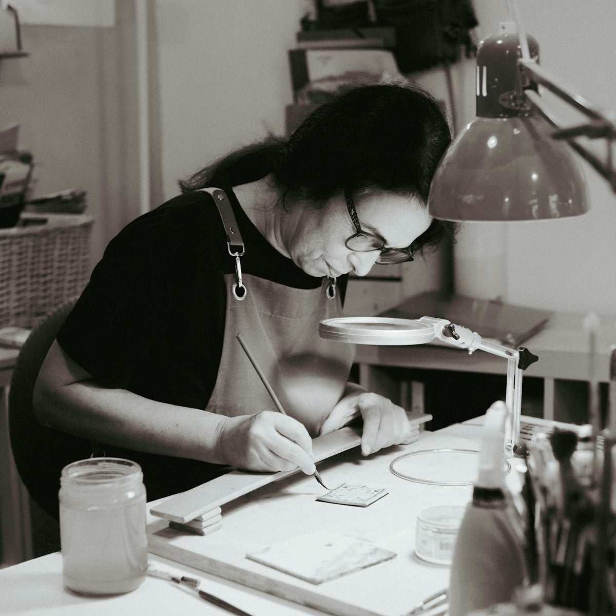 Woman painting at a desk with magnifying lamp.